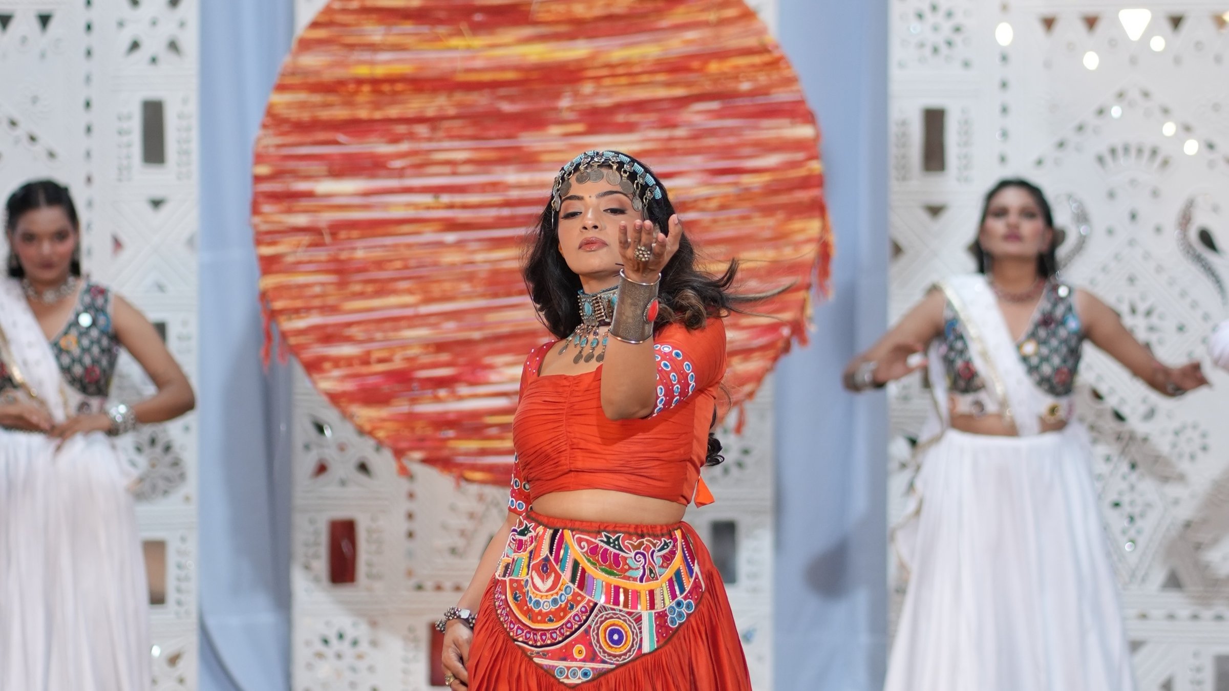 Pooja Kalyani in orange, centre stage between Garba dancers in white, a painted sun behind her