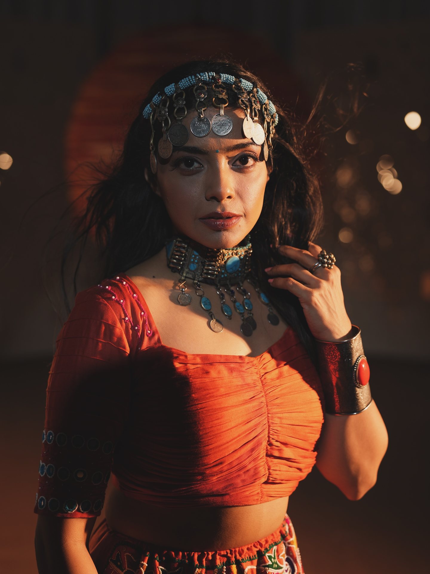 Pooja Kalyani looking into the camera in an orange chaniya choli, silver coin headpiece and turquoise necklace, lit by warm stage light